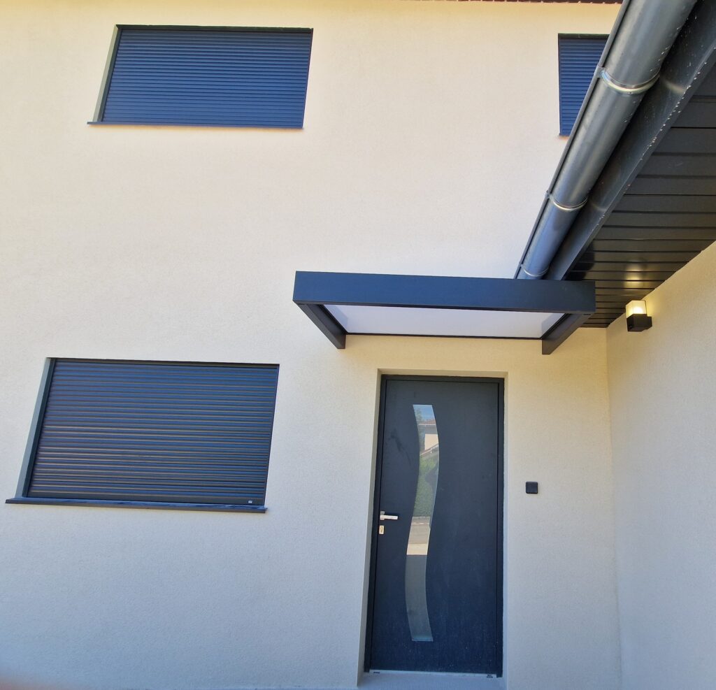 Façade de maison moderne crème avec porte d'entrée anthracite, stores roulants bleus fermés et auvent noir et blanc.
