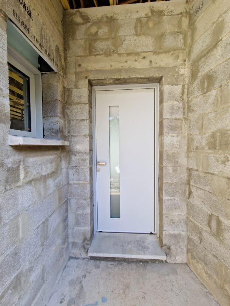 Porte d'entrée blanche neuve avec longue vitre verticale, encadrée par des murs en parpaings bruts et un seuil en béton. Une fenêtre est visible à gauche.