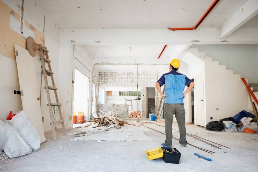 Homme en casque jaune et uniforme observe un vaste chantier de rénovation intérieur avec murs bruts, débris de bois, outils et matériaux.