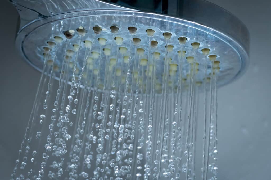 Pommeau de douche argenté sous l'eau, montrant des gouttes et filets d'eau limpides. Des dépôts de calcaire sont visibles sur les buses.