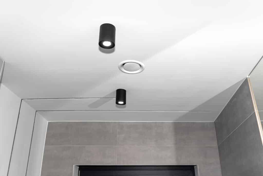 Éclairage moderne : Spots de plafond et carrelage gris Plafond blanc moderne avec deux spots noirs cylindriques allumés et une aération ronde. Murs carrelés gris, ambiance minimaliste.