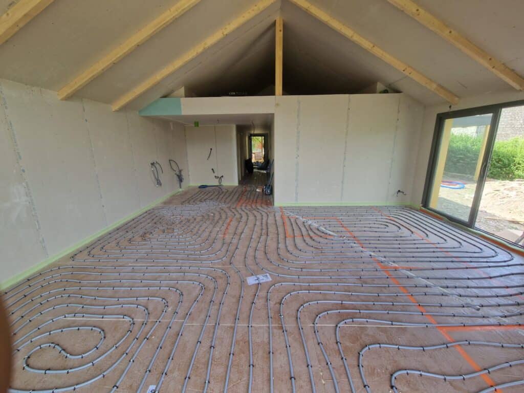 Chauffage au sol : pose des tuyaux en rénovation Installation de chauffage au sol avec tuyaux gris disposés en serpentin sur un plancher de construction, murs en plaques de plâtre.