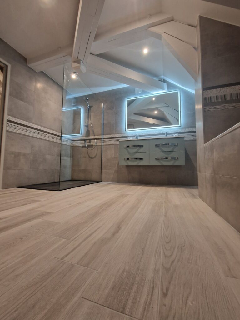 Salle de Bain Moderne Combles : Design Carrelage et Douche Salle de bain moderne sous combles avec carrelage gris, douche à l'italienne vitrée, meuble-lavabo suspendu et miroir lumineux à LED.