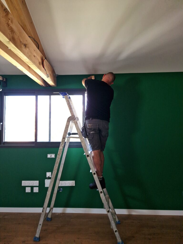 Installation électrique sur mur vert avec escabeau Un homme sur un escabeau fixe un élément au-dessus d'un mur vert et près d'une grande fenêtre lumineuse avec des poutres en bois.
