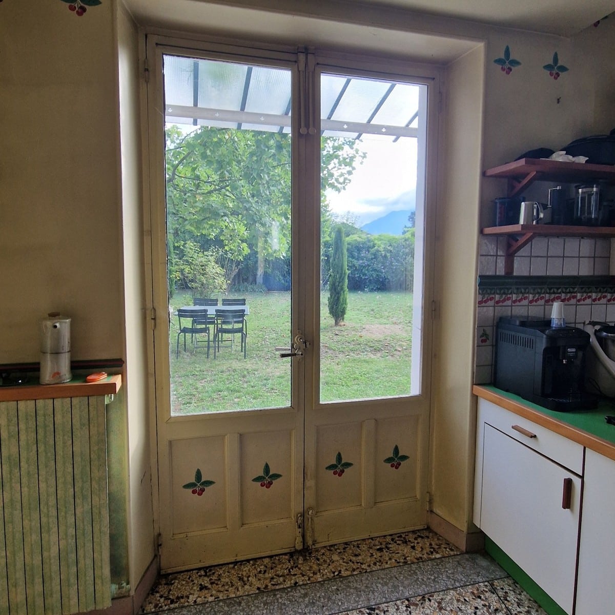 Porte-fenêtre de cuisine rustique ouvrant sur un jardin arboré avec table et chaises de terrasse.