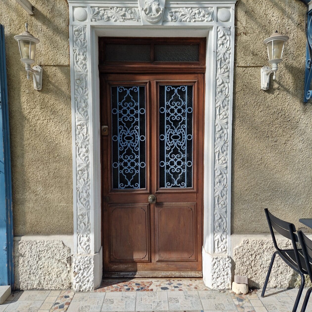 Porte en bois ancienne avec grilles bleues et encadrement blanc sculpté entre deux appliques murales.