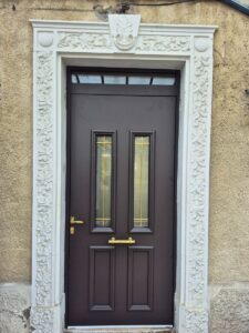 Porte d'entrée marron chocolat avec vitrage doré et encadrement en pierre blanche sculptée.
