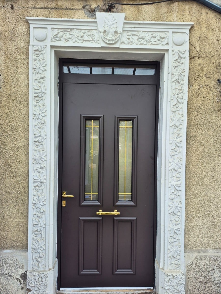 Porte d'entrée marron chocolat avec vitrage doré et encadrement en pierre blanche sculptée.