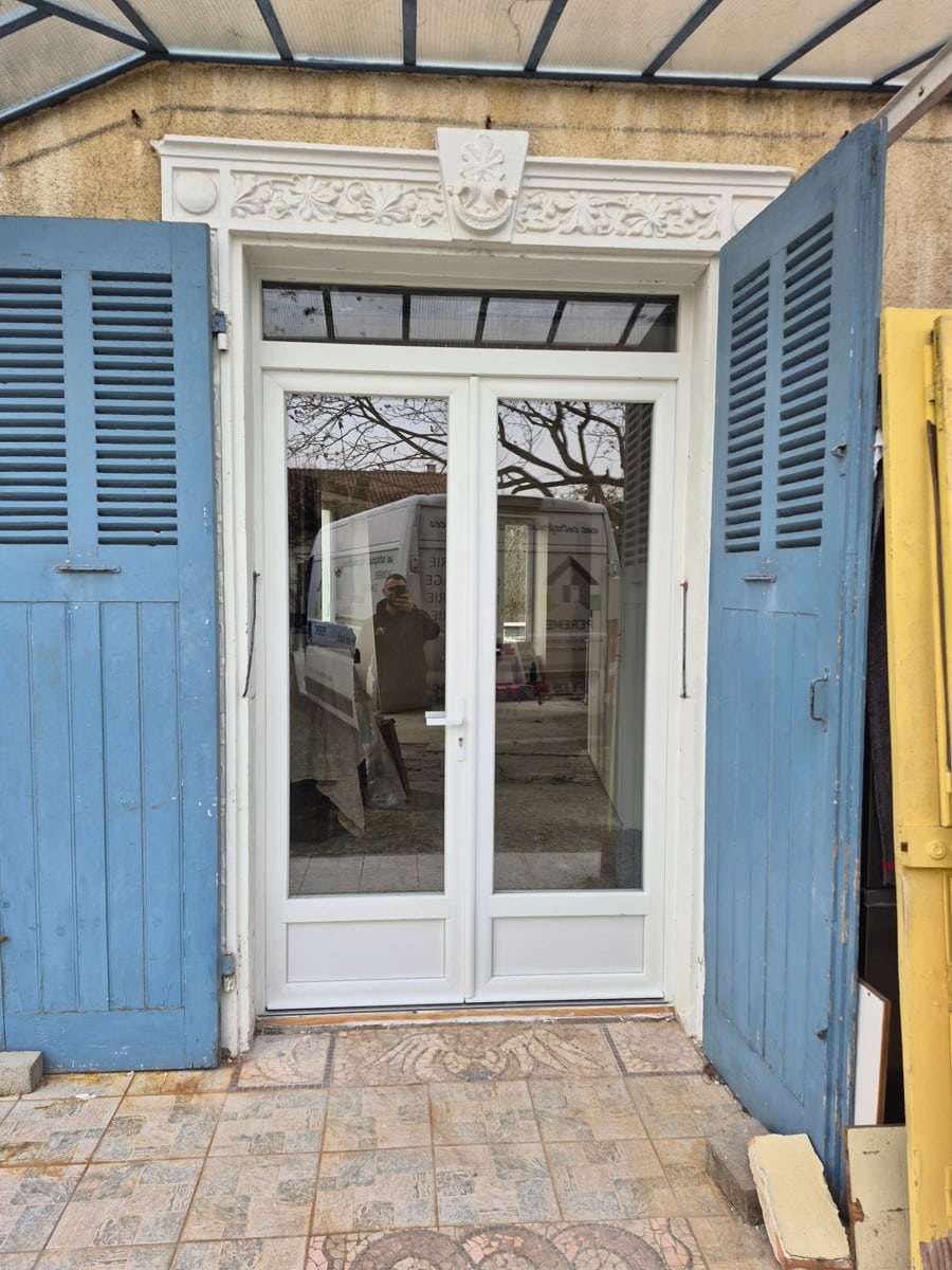 Porte-fenêtre blanche à double vitrage encadrée de volets bleus et d'un fronton sculpté.